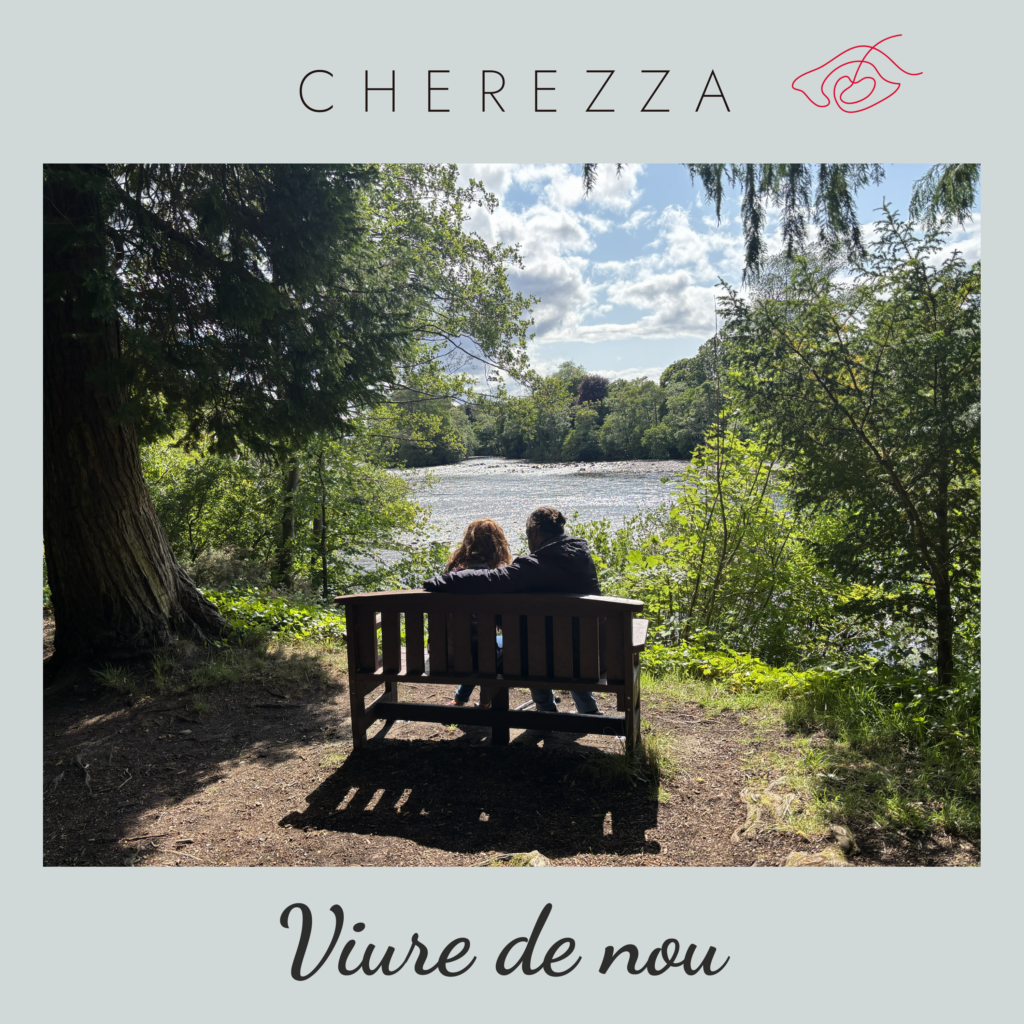 Una pareja, de espalda, sentada en un banco de madera, rodeado de vegetación, delante de un río. Arriba, el logo de Cherezza. Al pie, el título del single: Viure de nou.